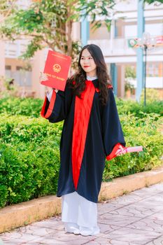 Smiling young woman in a graduation gown holding a diploma outdoors.