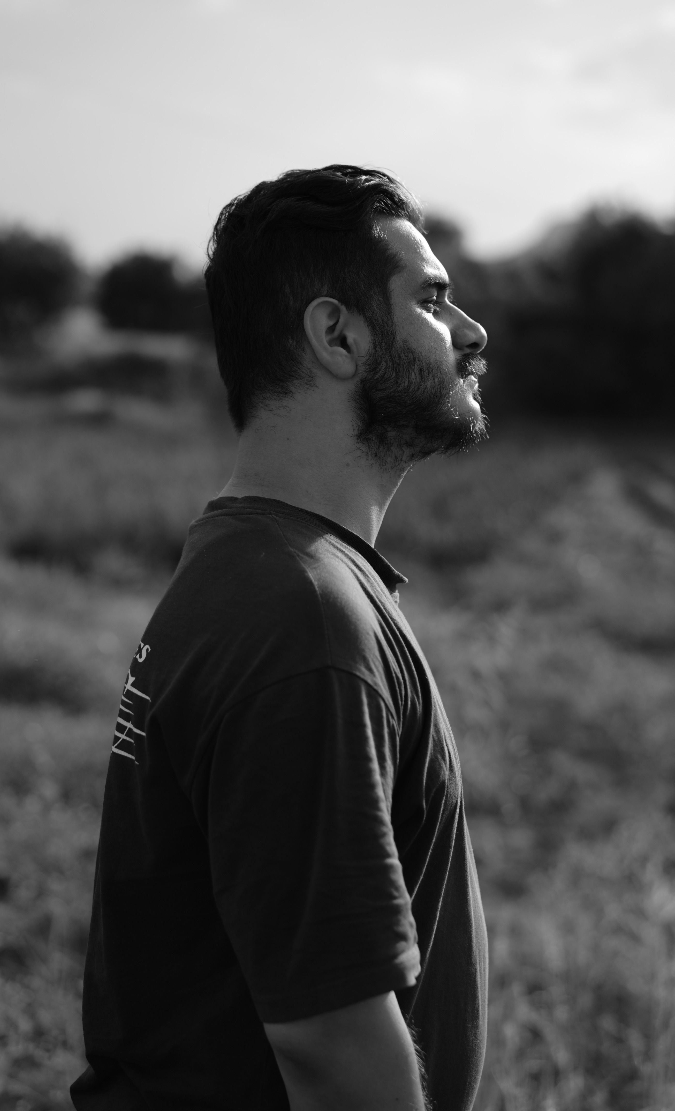 Artistic black and white profile portrait of a bearded man standing in a field.