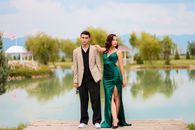 Stylish couple in formal attire posing by a tranquil lake with elegant gazebos and lush greenery.
