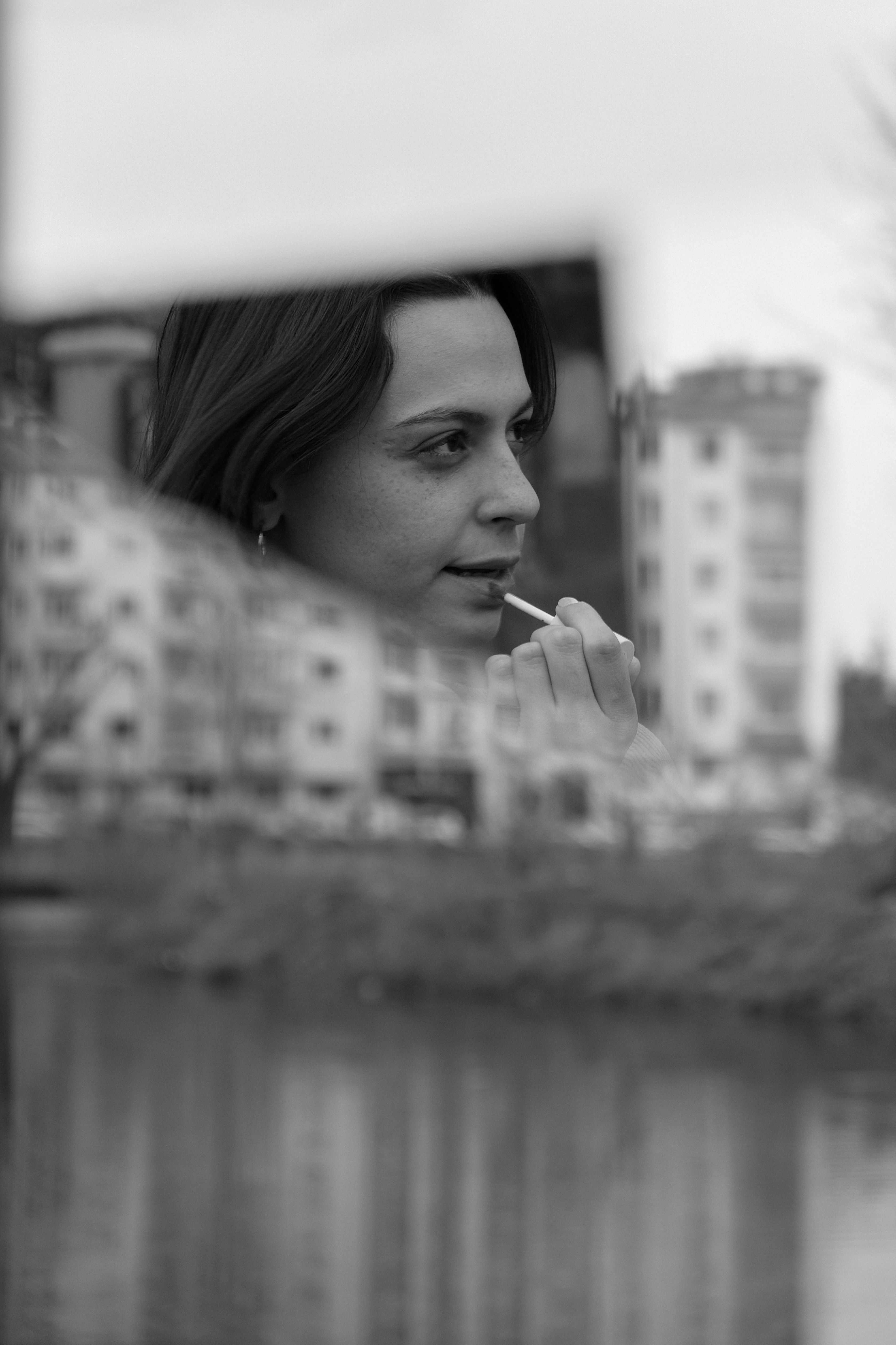 Black and white photo of a woman applying lipstick using a mirror reflection by the river