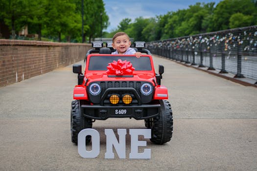 Happy toddler enjoying his first birthday in a red electric toy car outdoors.