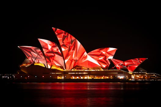 A vibrant night view of the Sydney Opera House lit in red patterns, reflecting on the water.