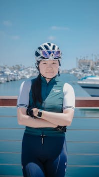 An Asian woman cyclist posing confidently by a marina in sportswear on a sunny day.
