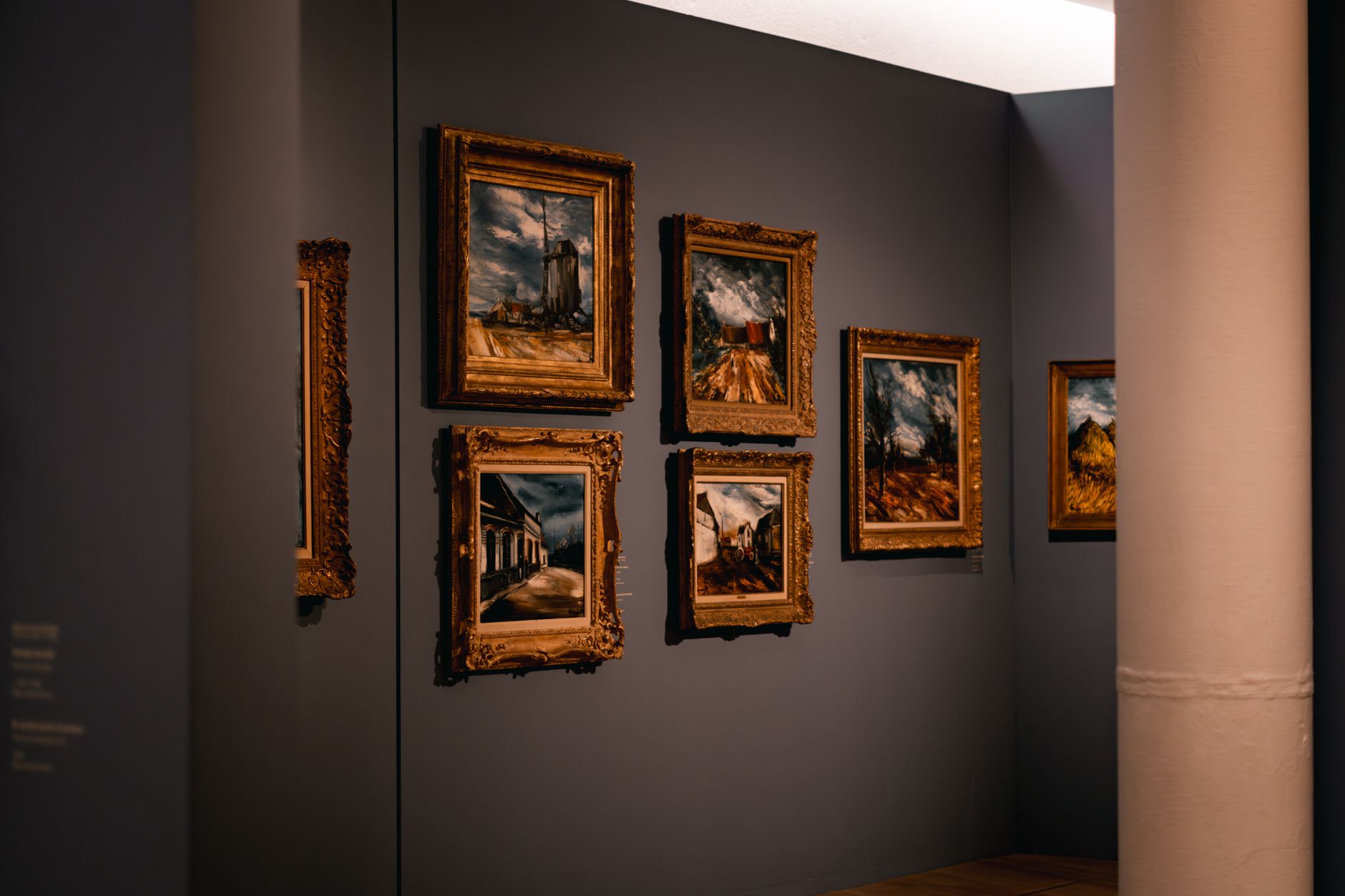 Modern bir sanat galerisinde beyaz duvarlara asılmış çerçeveli tabloların ve aydınlatmalı sergi alanının görünümü.
