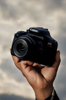 A close-up view of a DSLR camera being held aloft against a cloudy sky backdrop.
