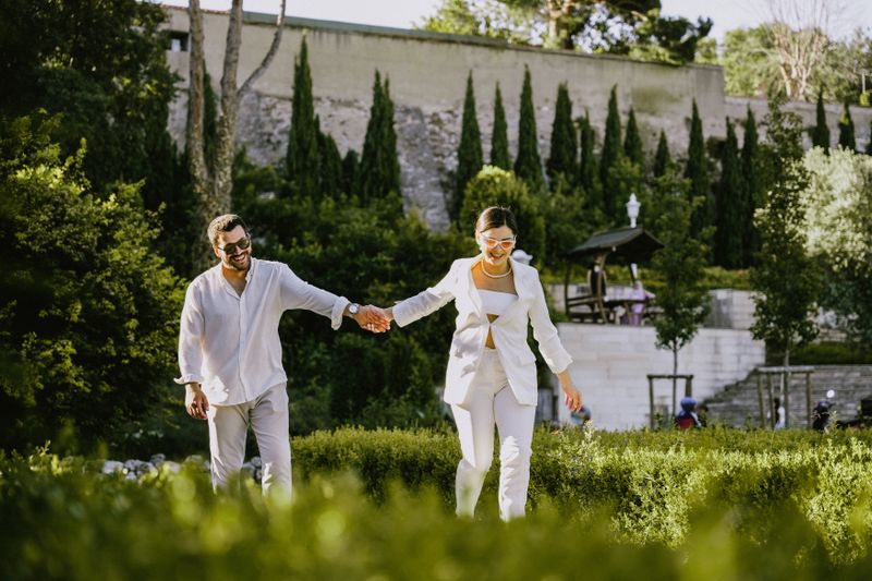 Chic couple holding hands and walking in a sunlit park, exuding joy and style.