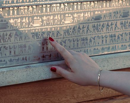 A woman's hand with red nails touching a metallic wall with hieroglyphs under soft lighting.