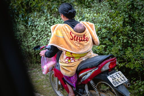 The woman wraps her child in a blanket on her back while riding a motorcycle.