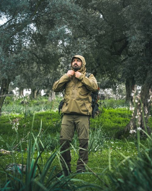 Free A man in a green jacket adjusting his hood while hiking in a lush, green forest setting. Stock Photo