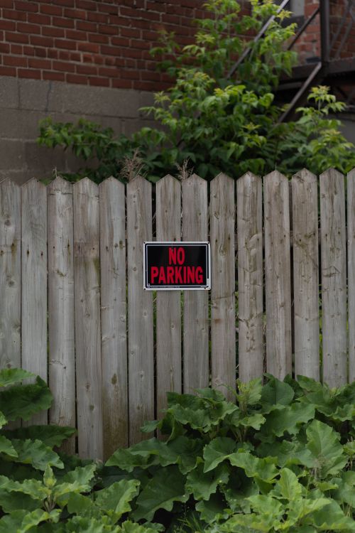 Free A vertical shot of a wooden fence with a no parking sign and lush greenery Stock Photo