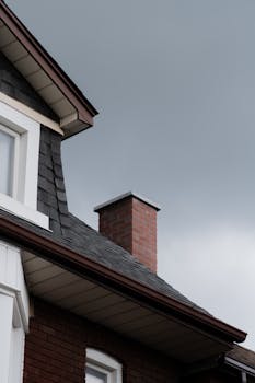 Close-up of a house roof with a brick chimney against a cloudy sky.