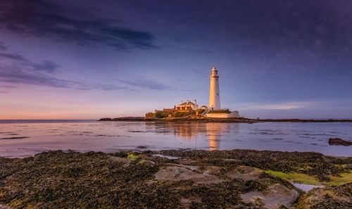 Free A picturesque lighthouse stands on a rocky shore, reflecting in calm waters at sunset. Stock Photo