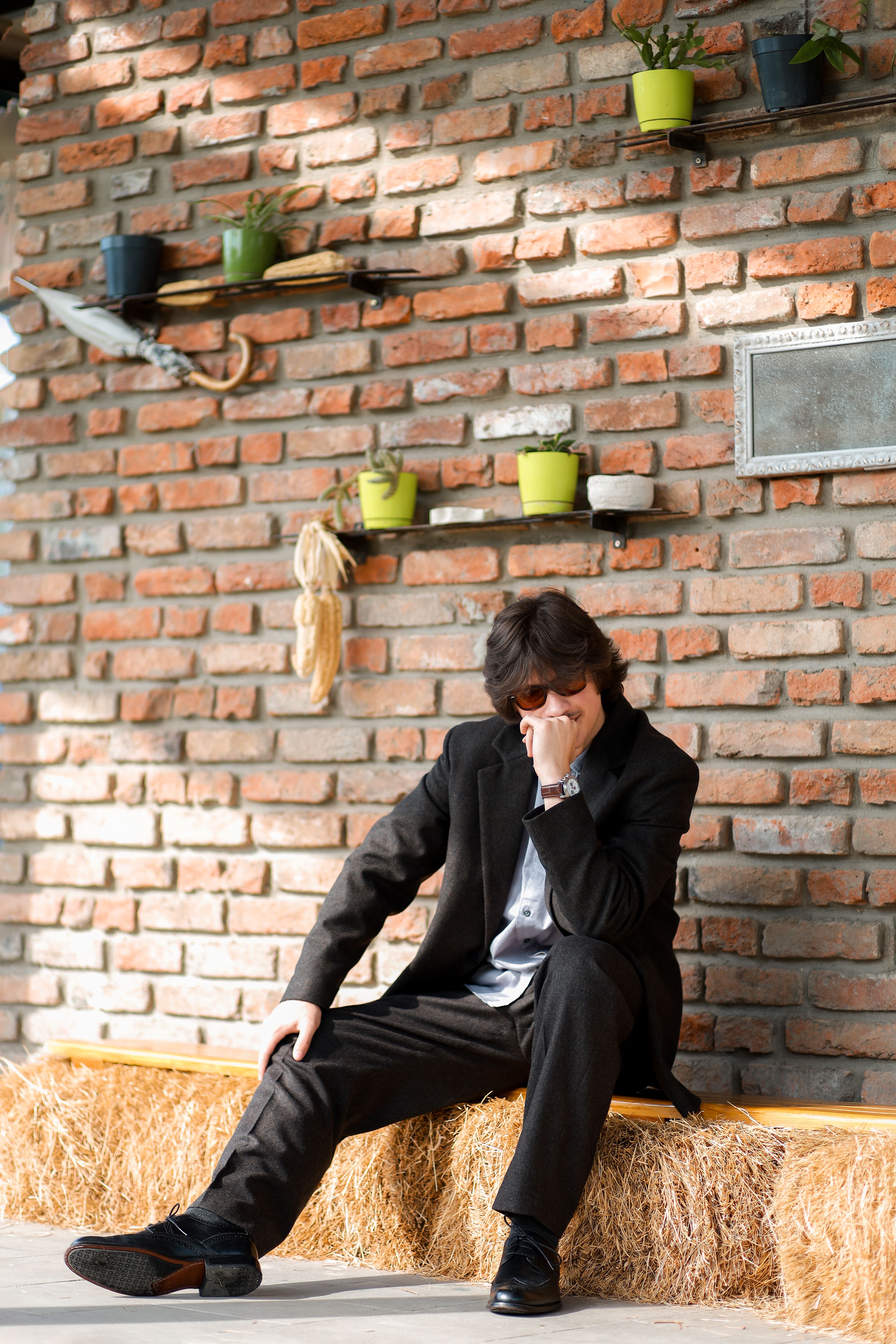 Man in suit with sunglasses sitting on hay bales against brick wall with flowerpots.