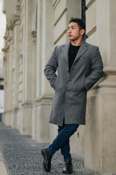 A fashionable man in a gray coat leaning against a building exterior in an urban setting.
