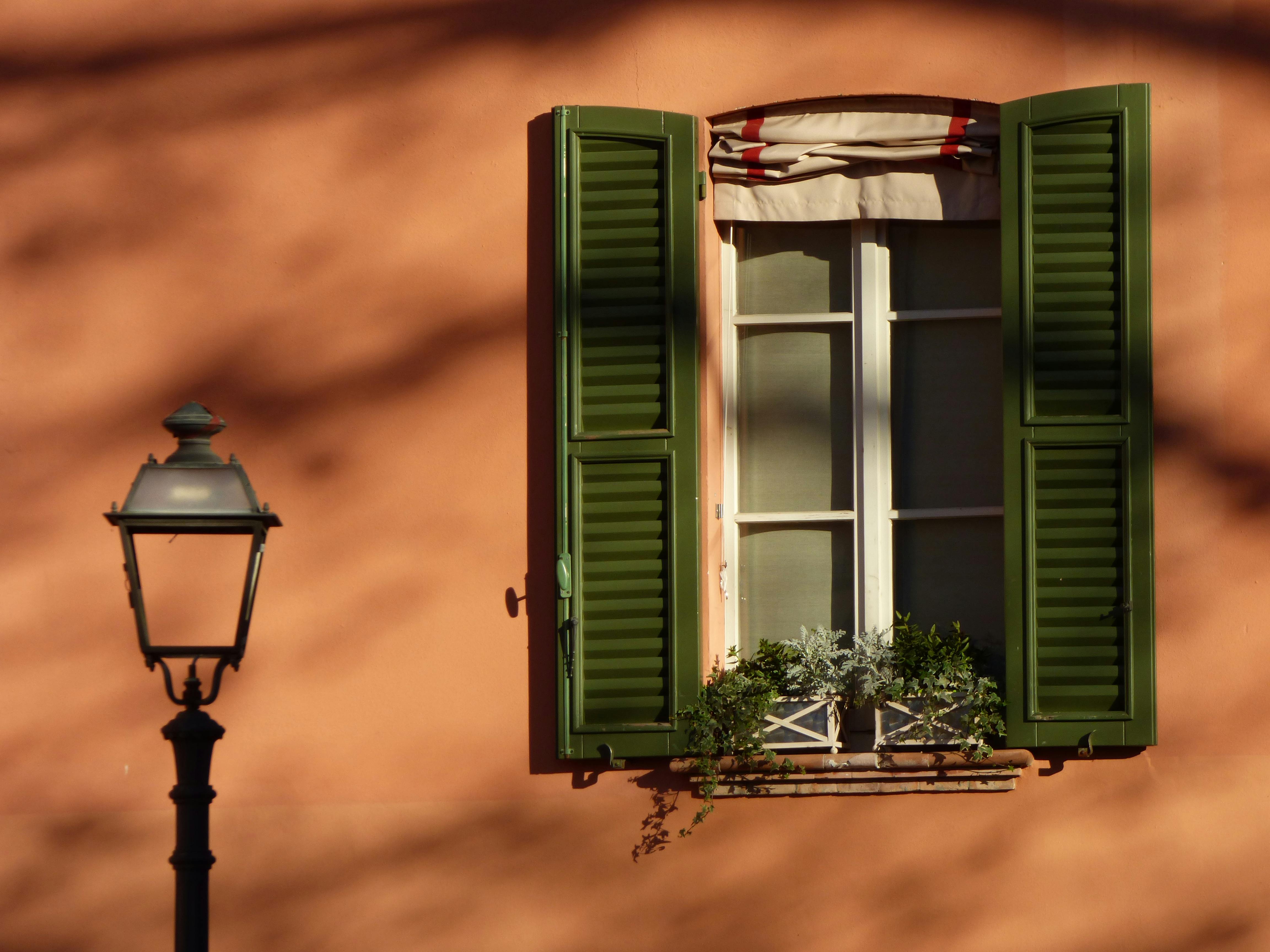 Sunlit window with green shutters and street lamp in Parma, Italy.