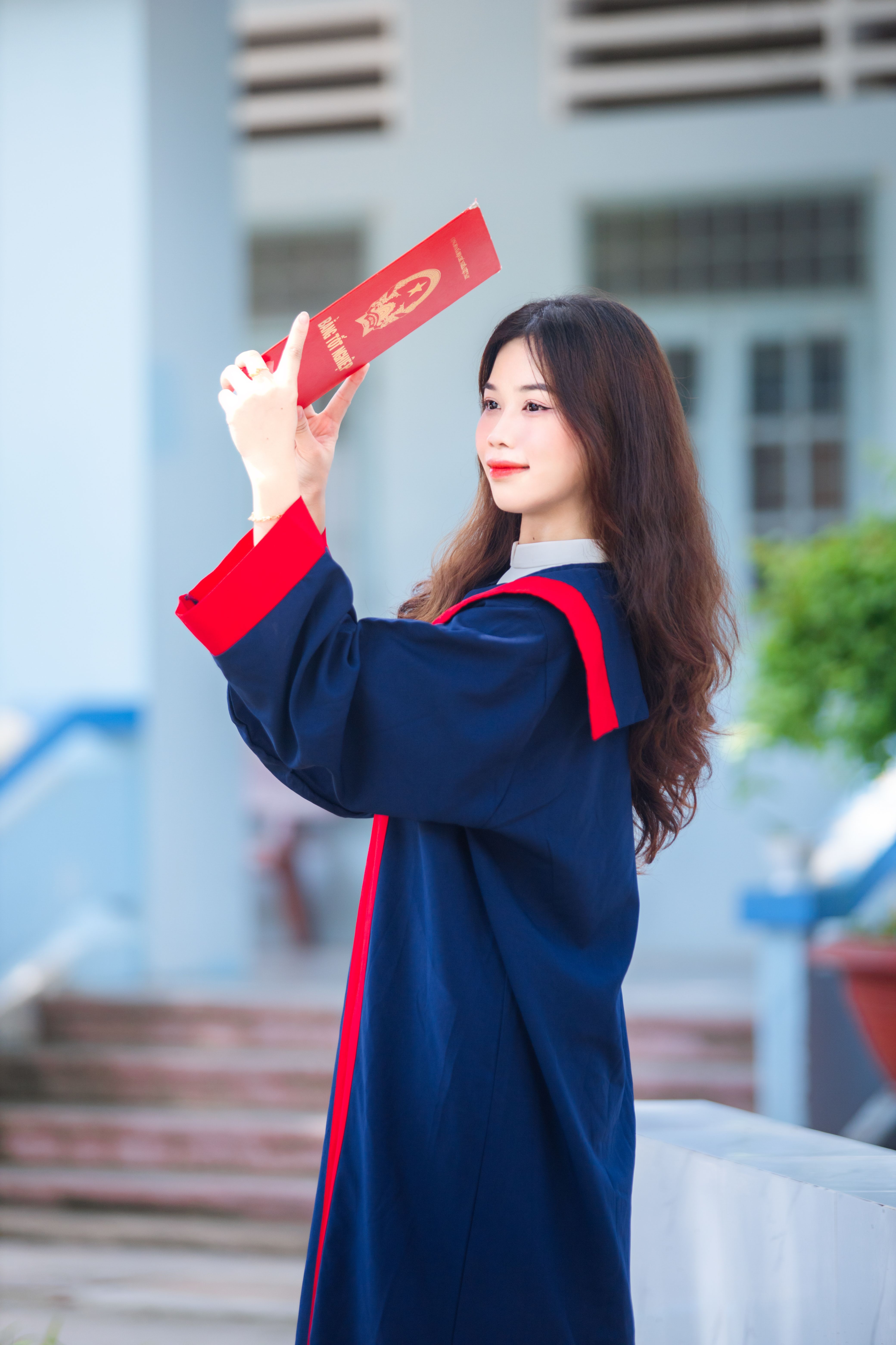 Asian woman in graduation gown holding diploma outdoors, celebrating academic achievement.