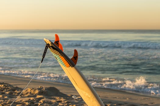 A surfboard planted in the sand by the ocean waves at sunrise, evoking a sense of adventure.
