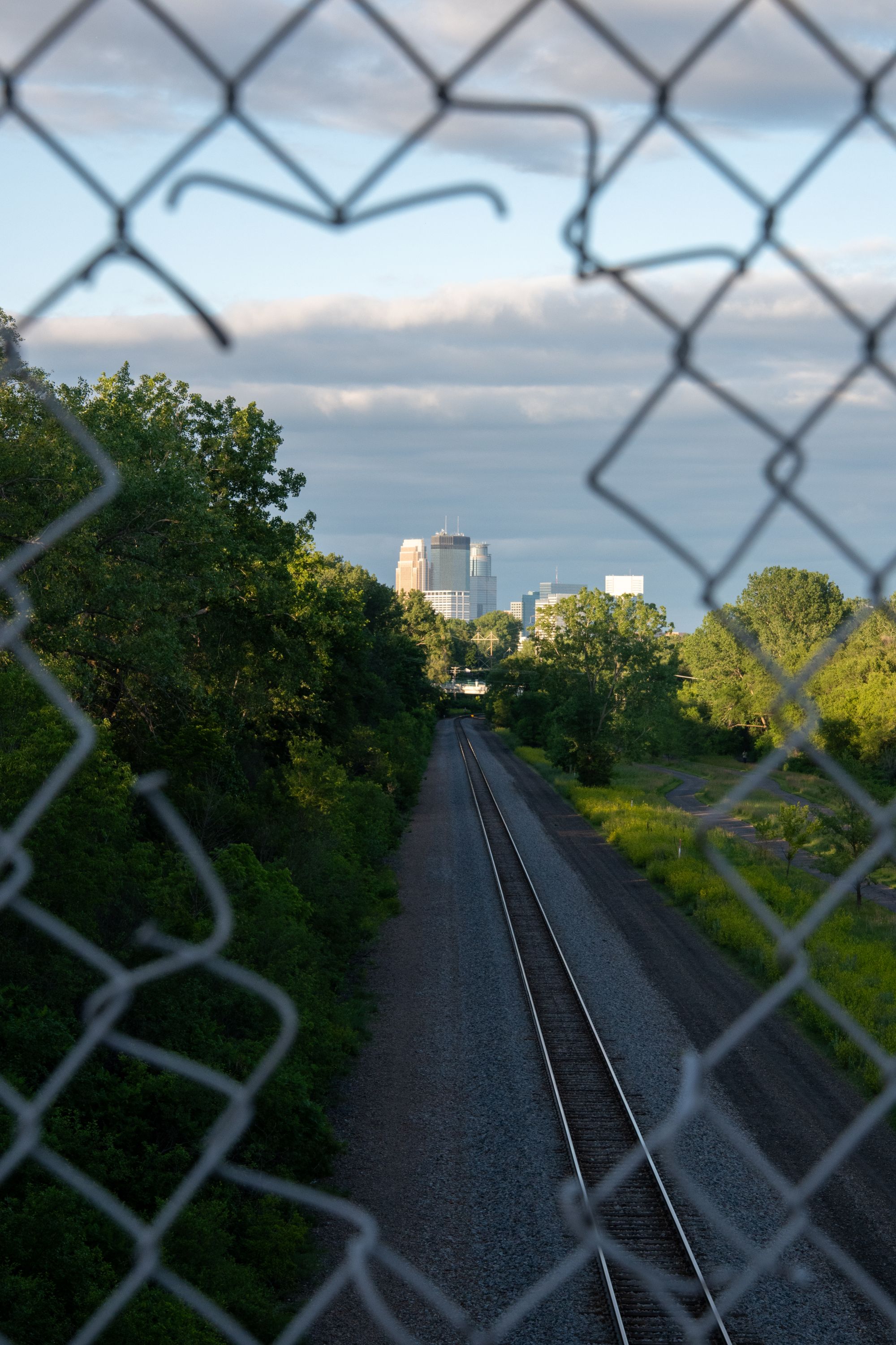 A railway track leading toward a city skyline, seen through a gap in a chain-link fence