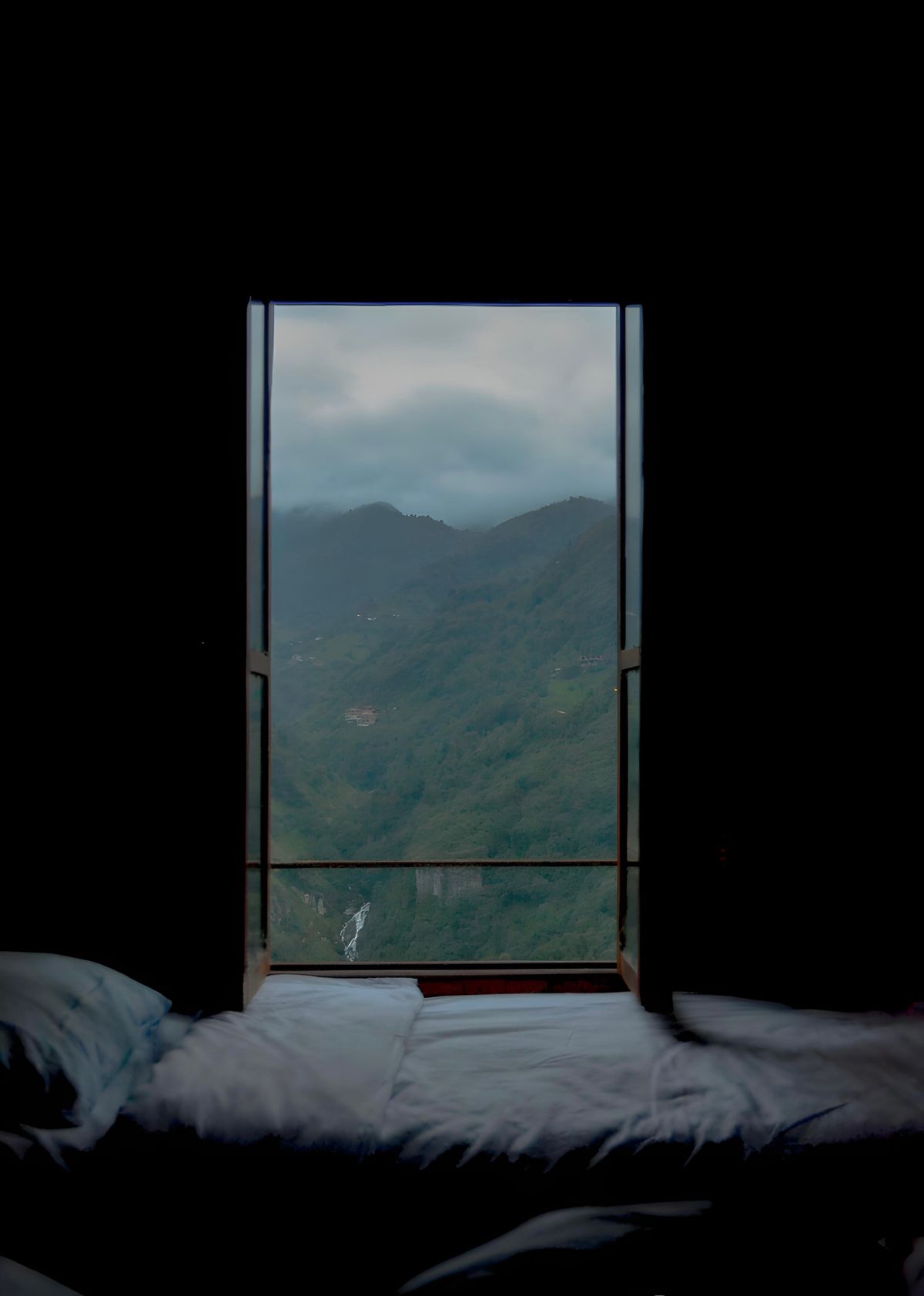A plain bed in a dark room, the window open to forested ridges and cloud on the far hillside