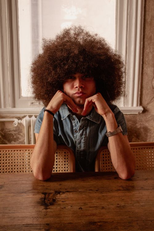 Free Young man with curly hair resting chin on hands at wood table in fashion setting. Stock Photo