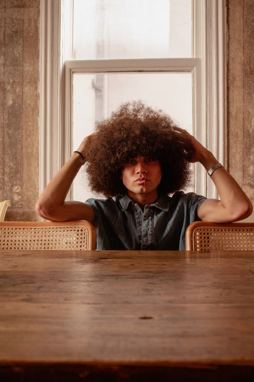 Free Artistic portrait of a man with curly afro hair sitting indoors by a window. Stock Photo