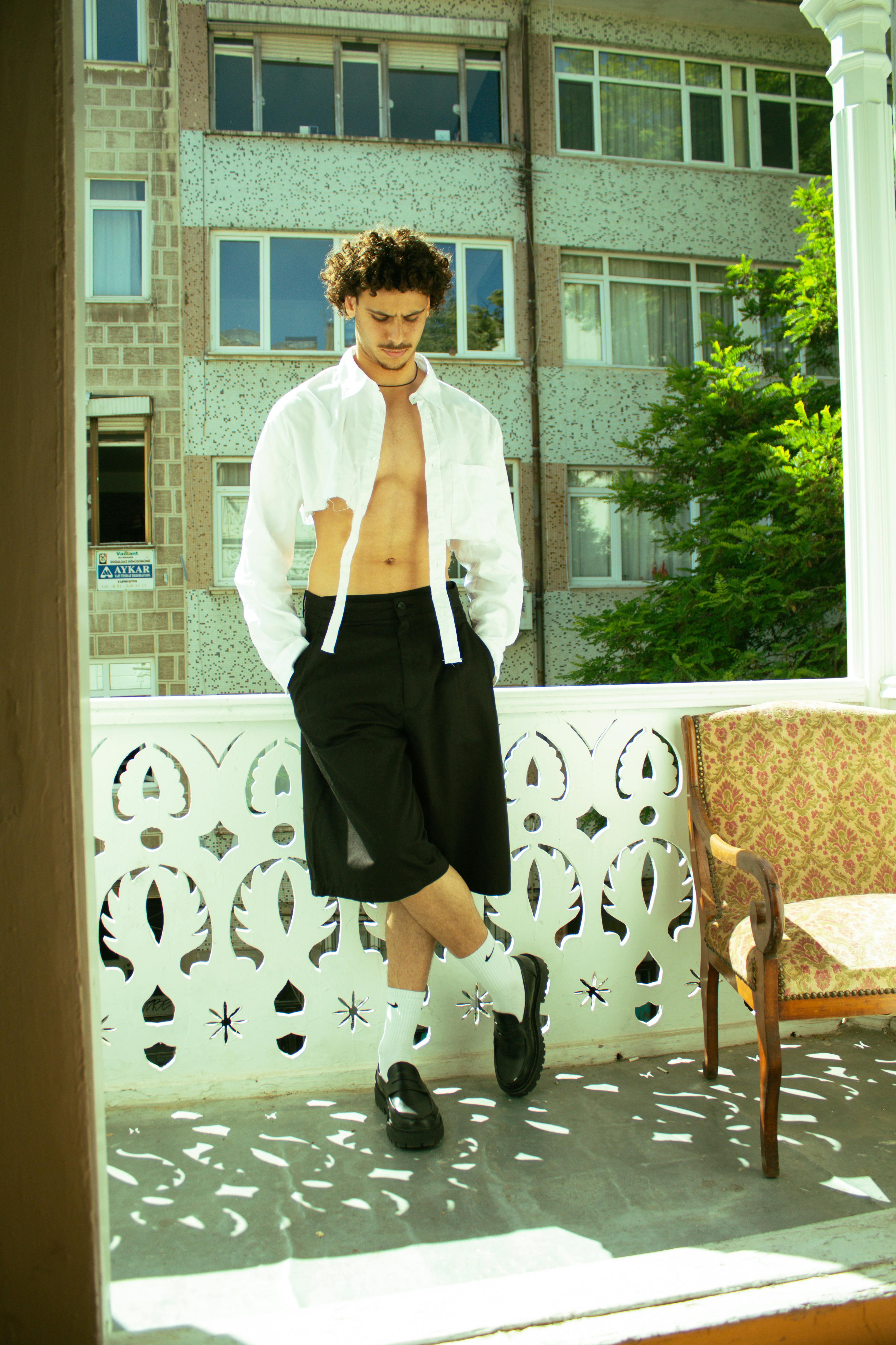 Stylish male model posing on a sunlit balcony in urban setting, wearing modern casual clothing.
