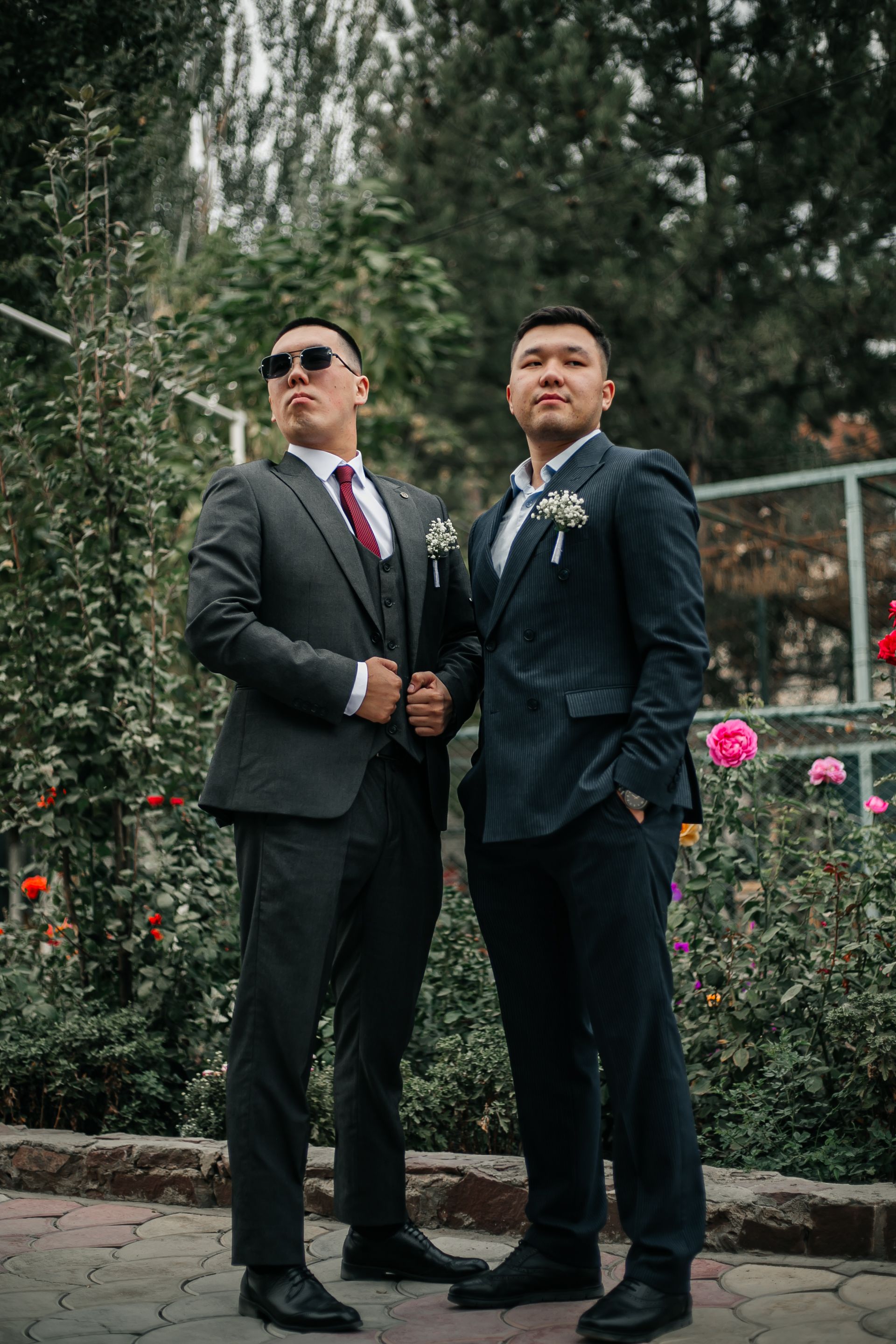 Two men in suits posing confidently in a garden with blooming flowers in Tokmak, Kyrgyzstan.