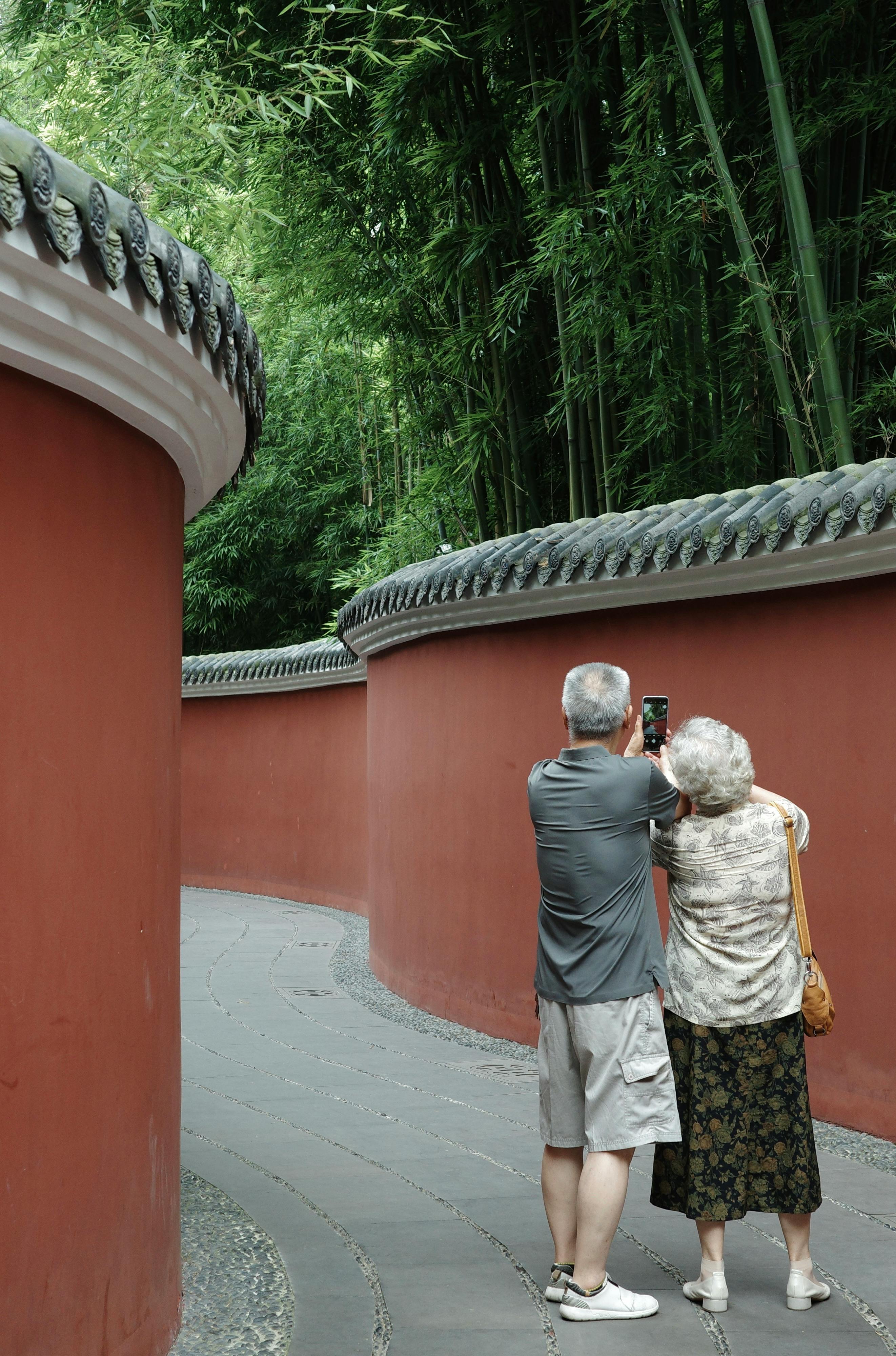 Senior couple taking a selfie along a red wall pathway surrounded by bamboo.