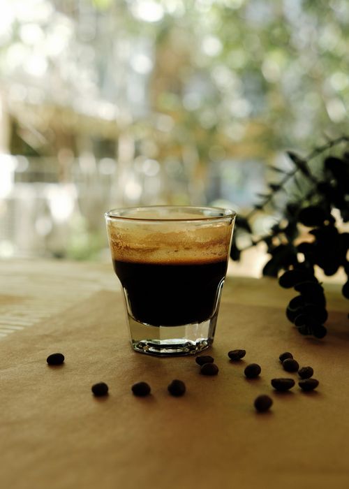Free Shot of an espresso in a glass on a wooden table with scattered coffee beans and blurred background. Stock Photo