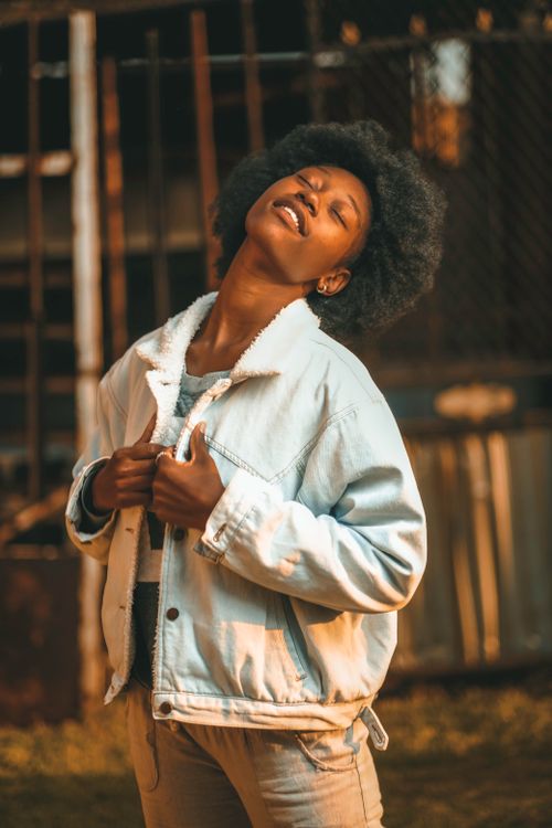 Free Young woman in denim jacket poses outdoors in urban South African neighborhood, basking in sunlight. Stock Photo