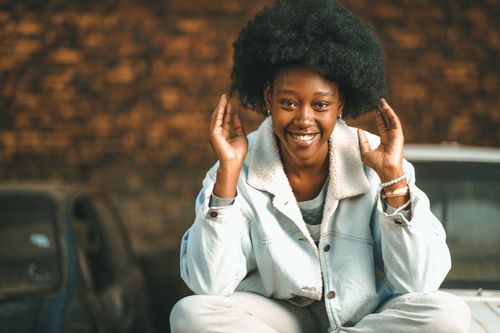 Free Happy young woman with afro hairstyle sitting outdoors, smiling and gesturing playfully. Stock Photo