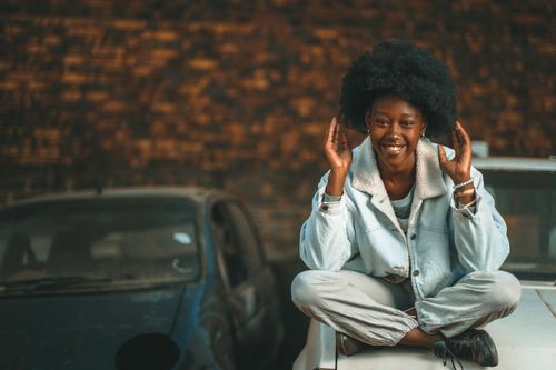 Free Portrait of a young woman with an afro hairstyle sitting on a car and smiling outdoors. Stock Photo