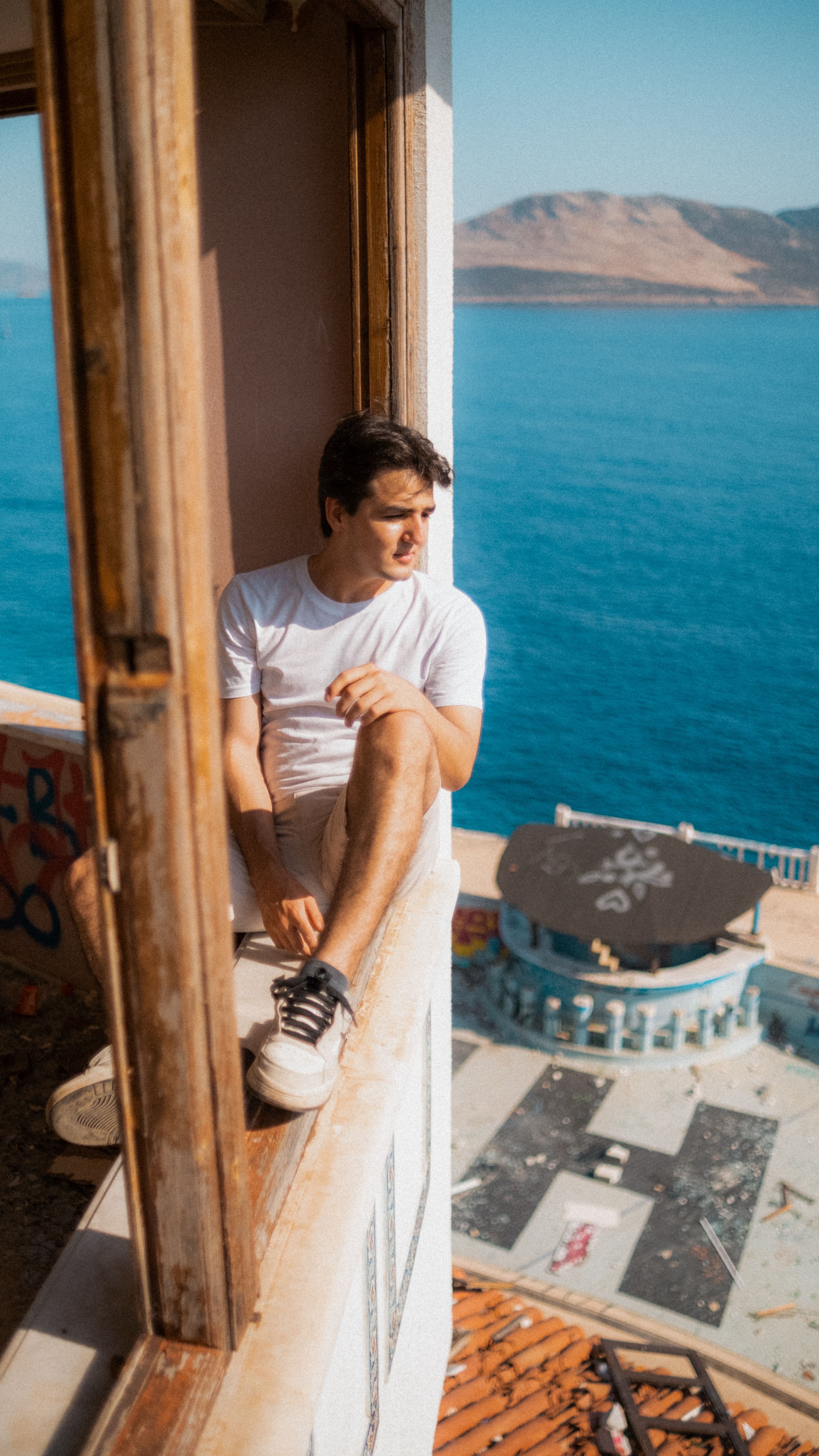 A young man in a white t-shirt sitting on a window sill overlooking the sea, conveying a sense of tranquility.