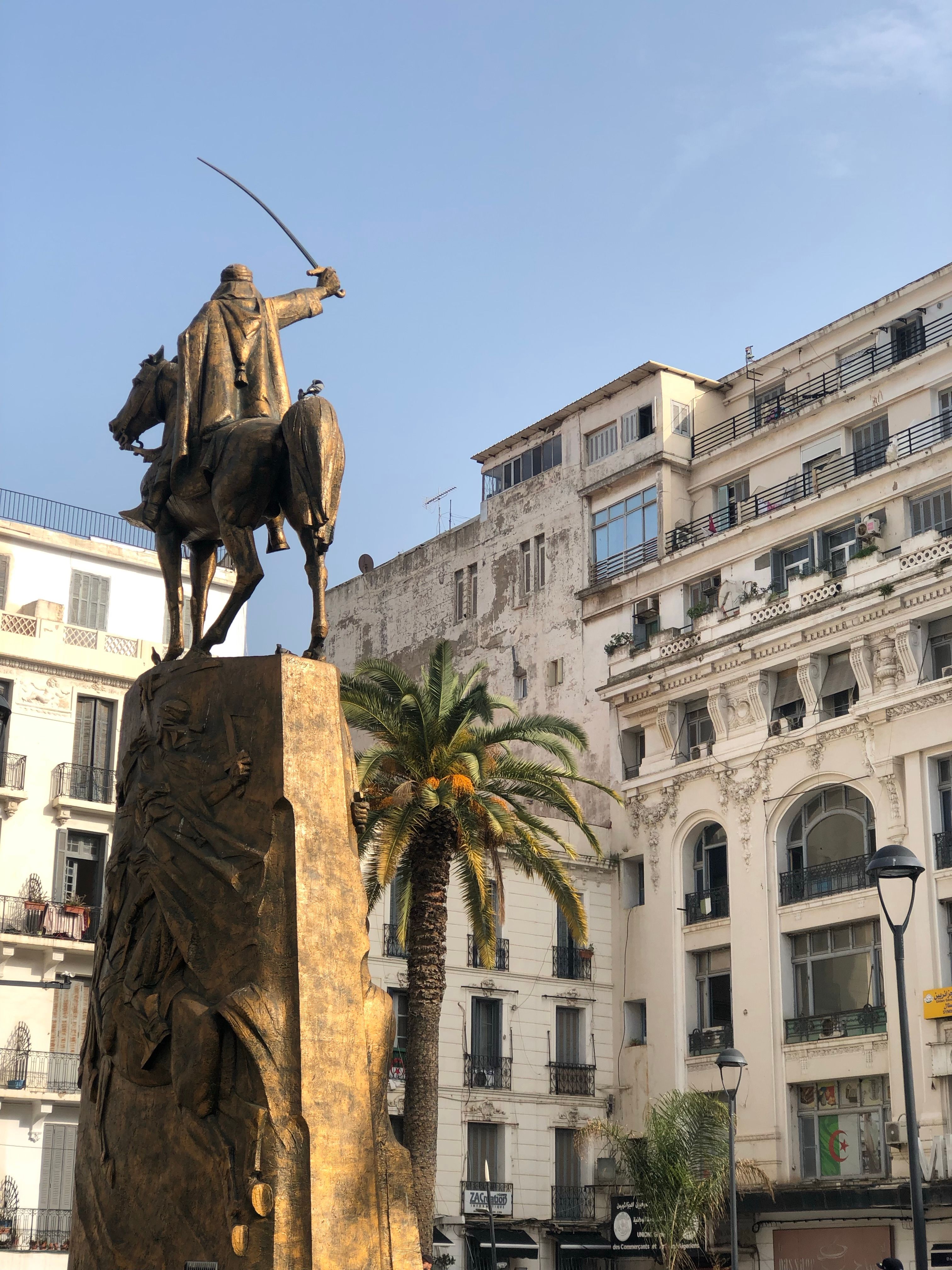 A striking view of the Emir Abdelkader statue amidst Algiers' urban architecture and palm trees.