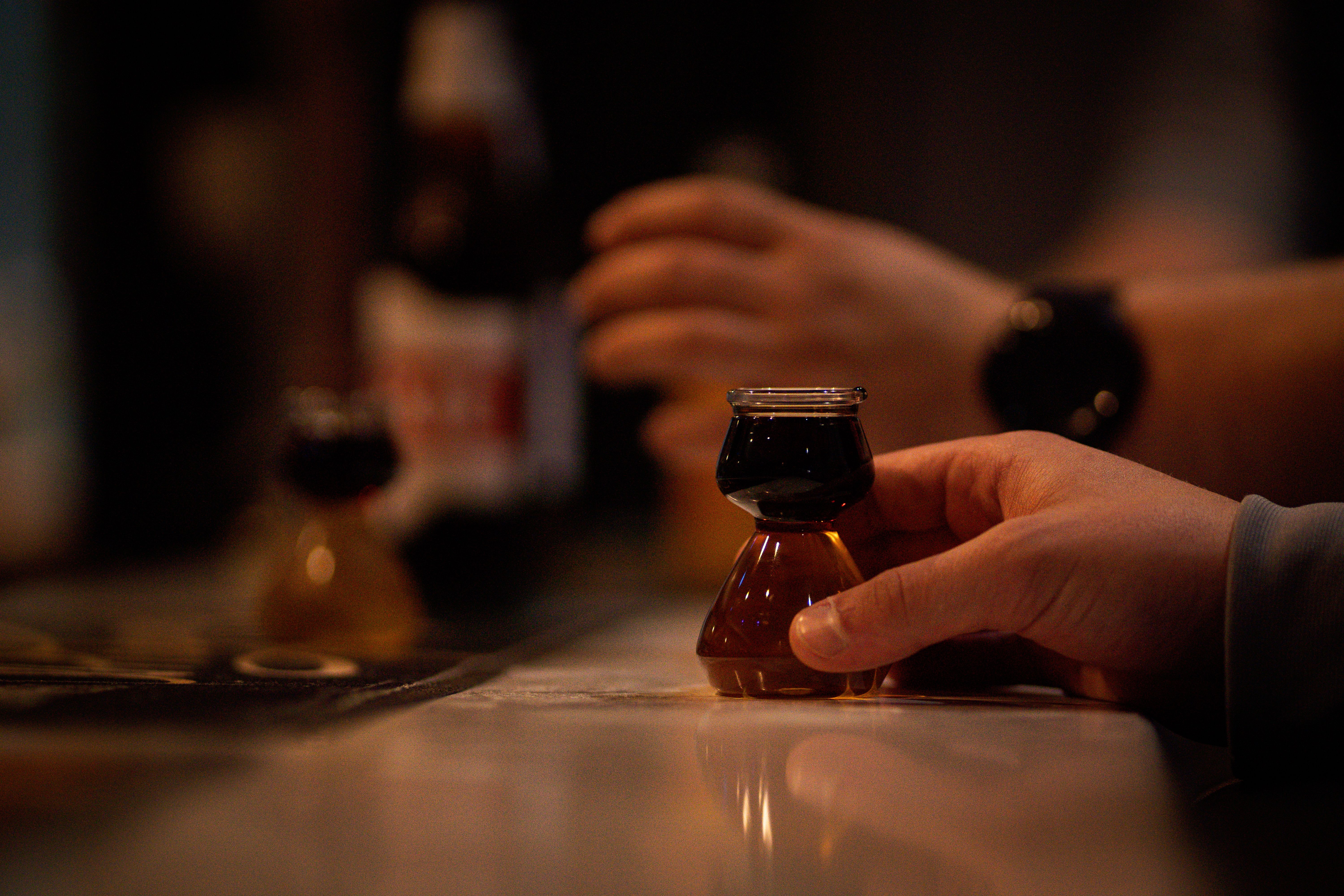 A close-up of a person holding a small glass of dark liquor at a bar counter, creating a warm and inviting mood.