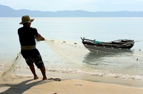 Free A fisherman on a beach casts his net near a fishing boat at sea, showcasing traditional fishing methods. Stock Photo