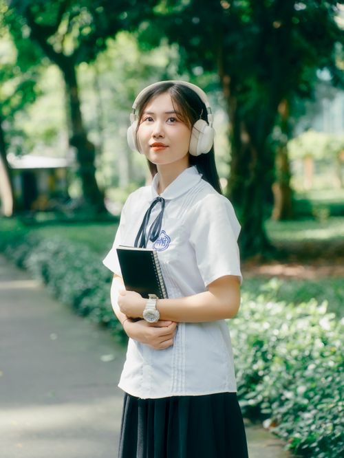 Free Asian woman standing in a sunny park, holding a notebook and wearing headphones. Stock Photo