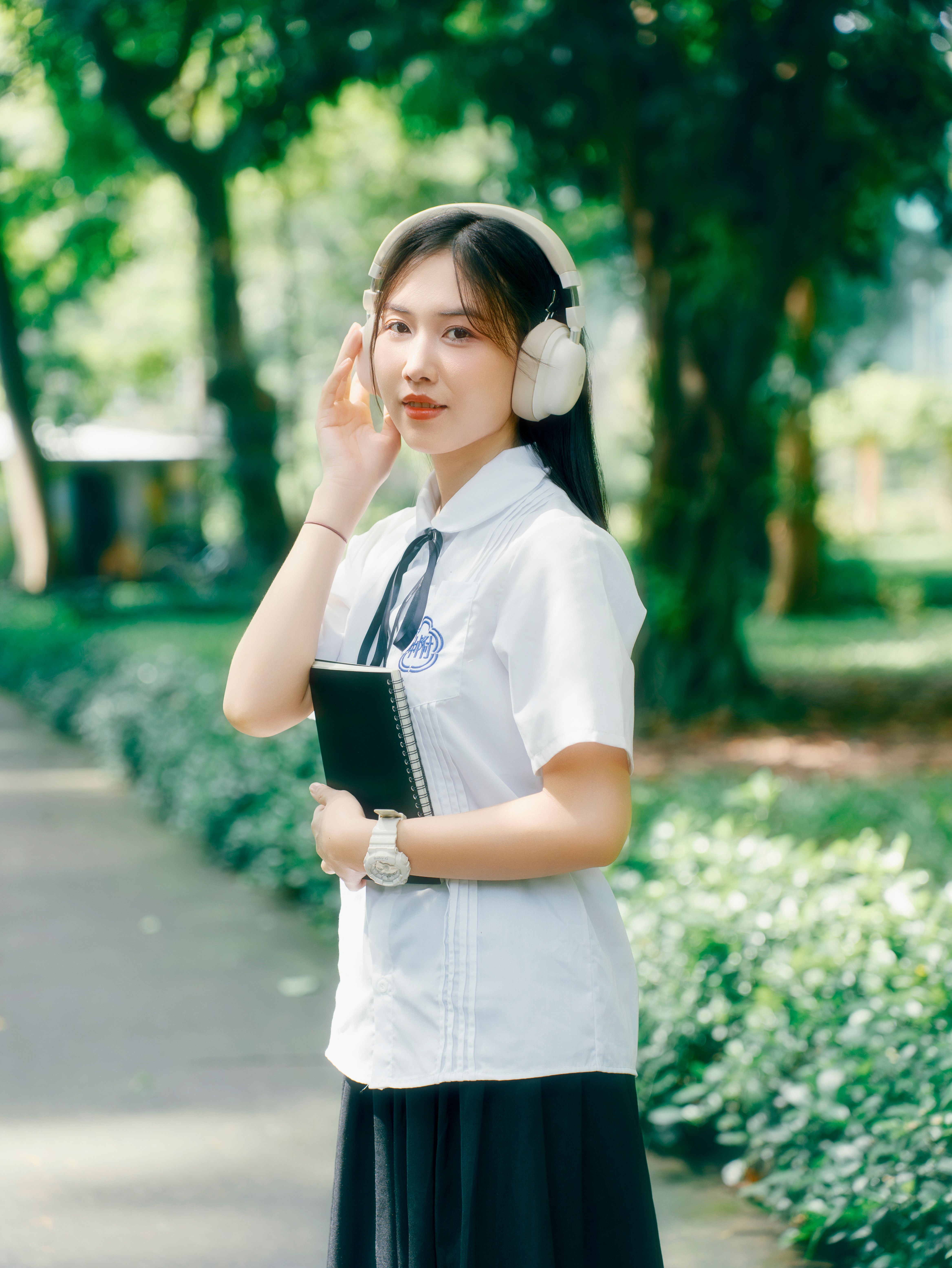 Asian woman in uniform with headphones, enjoying music in a lush park setting.