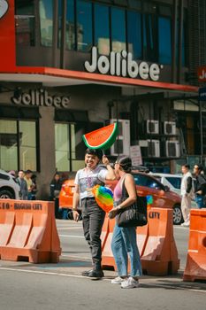 Two people with a watermelon umbrella in front of a Jollibee store, vibrant city scene.