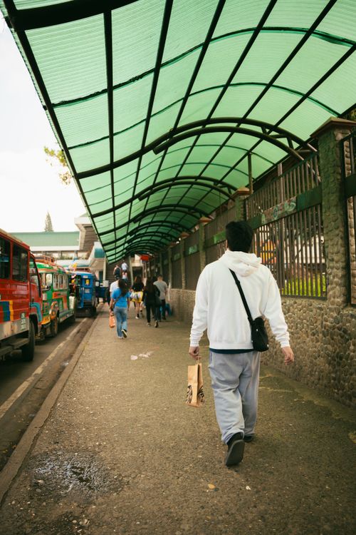 Free A man in a white hoodie walks down an urban sidewalk under a green canopy. Stock Photo