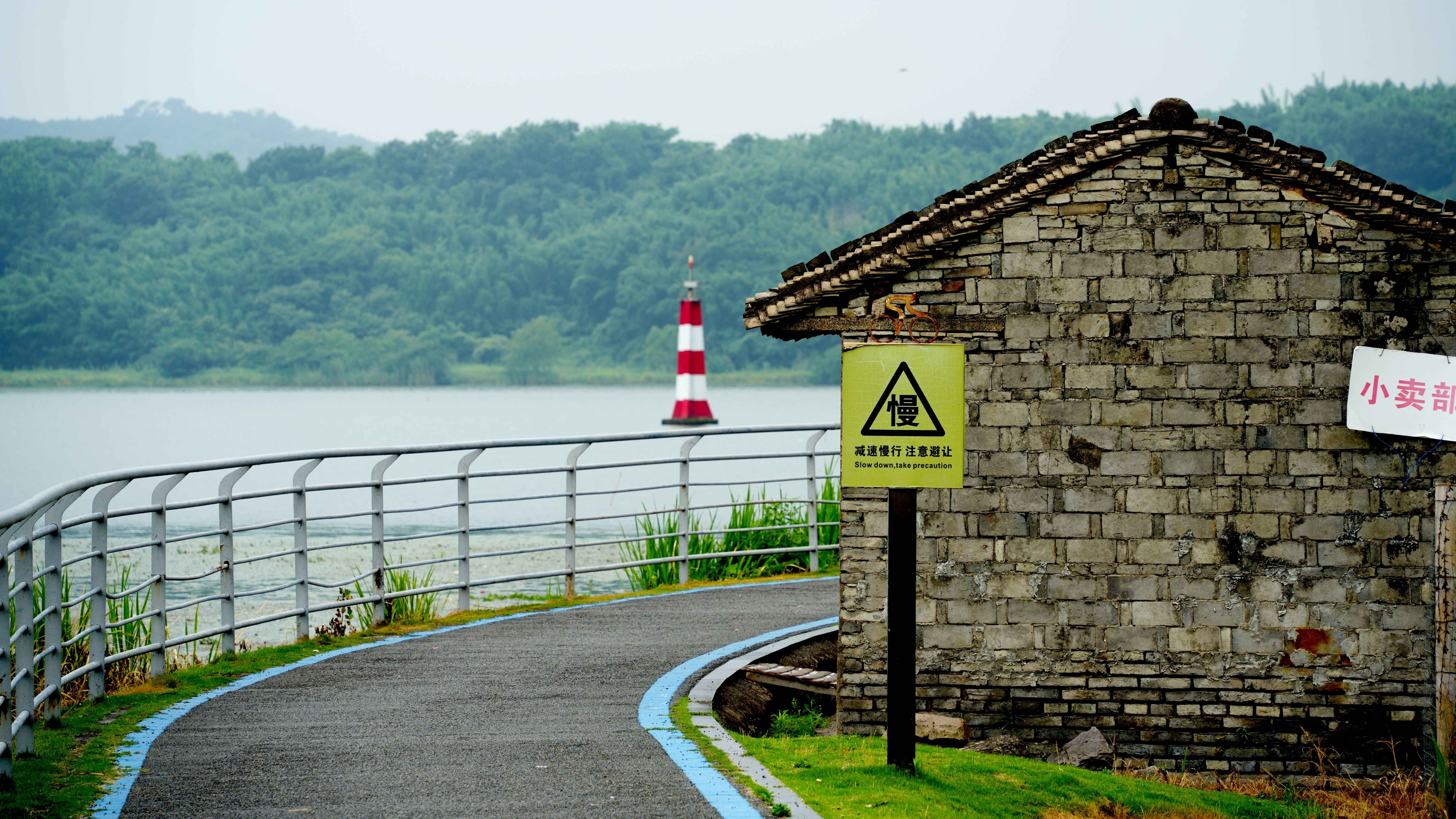 Scenic view of a riverside pathway with a brick building and distant lighthouse amidst forested backdrop.