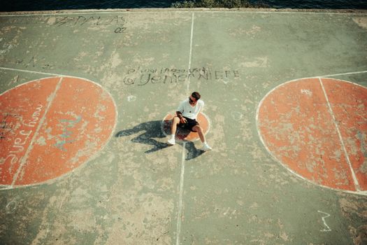 A man sitting on a weathered basketball court, viewed from above, in an urban setting.
