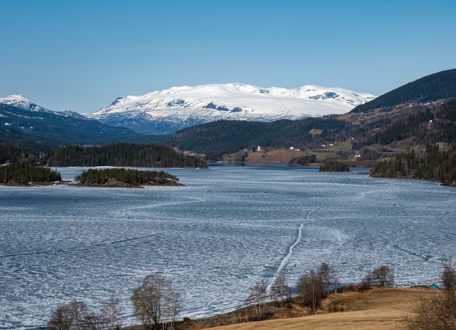 Scenic winter view of frozen lake and snow-capped mountains in Aurdal, Norway, under clear blue sky.