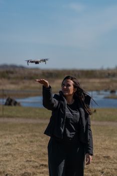 A woman in a black jacket flies a drone in a wide open field on a sunny day.
