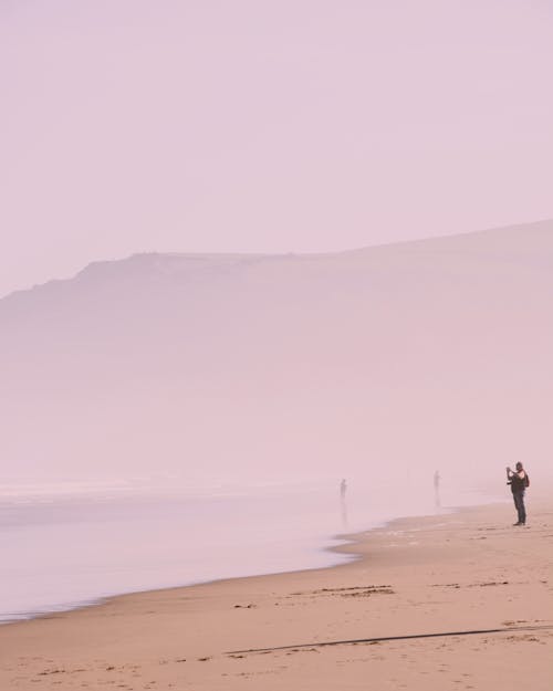Person taking picture of calm sea in hazy veil while standing on quiet sandy beach on foggy day