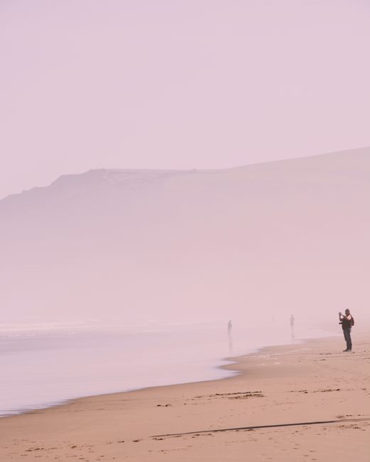 Person taking picture of calm sea in hazy veil while standing on quiet sandy beach on foggy day