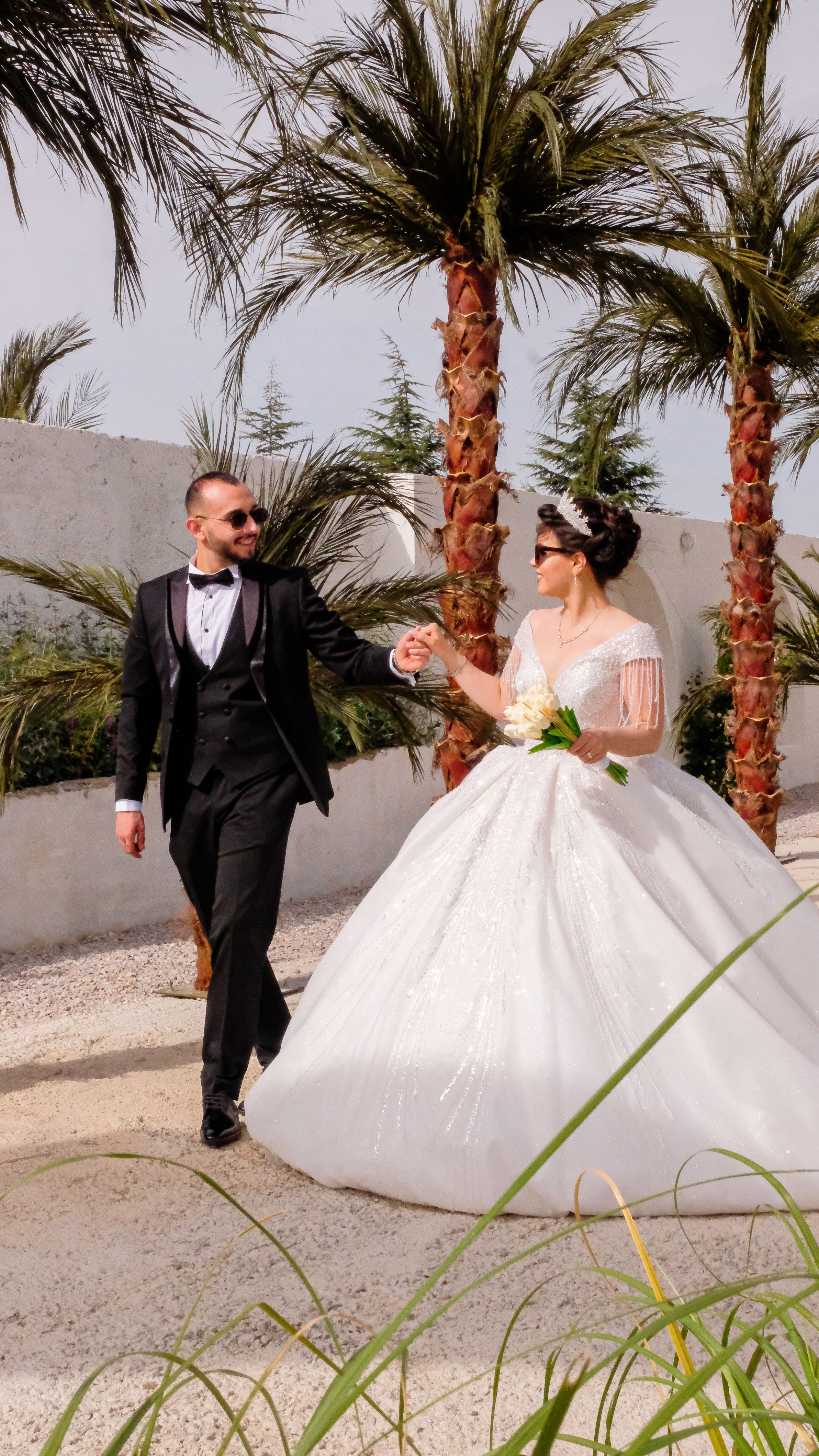 Bride and groom walking hand in hand outdoors amid palm trees, capturing a joyful wedding moment.