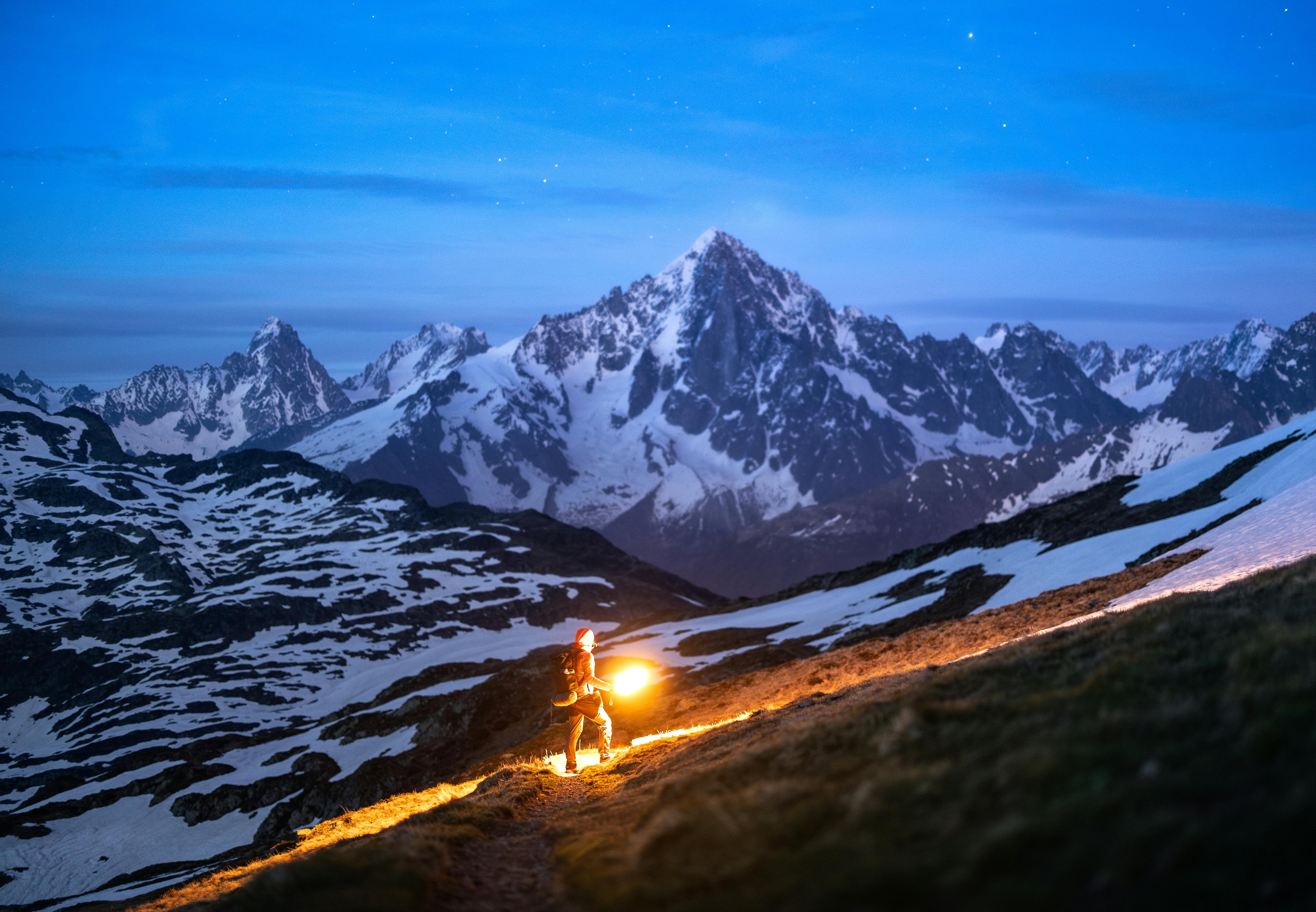 A lone hiker illuminates the snowy Alps at night, a dramatic adventure scene.