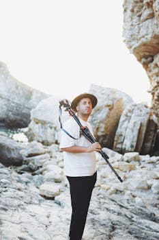 Photographer standing with a tripod in a rocky landscape, wearing a hat.