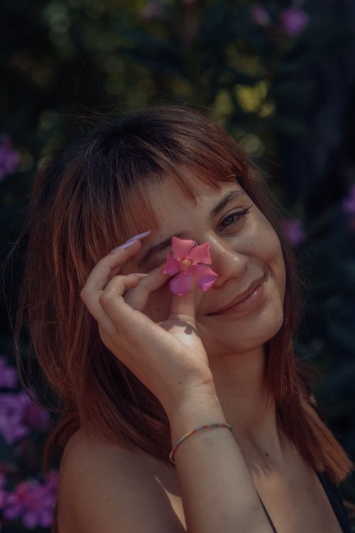 Free Young woman with bangs smiling, holding a pink flower near her face outdoors. Stock Photo